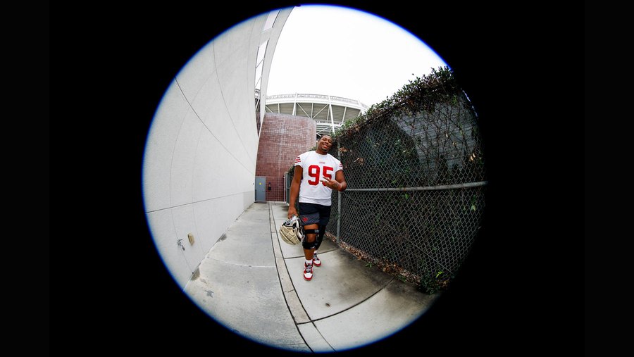 Alfred Collins, number 95, in a white 49ers jersey with red numerals, walking a fenced corridor toward the field carrying his helmet, shot through a fisheye lens. This is a 2025 club practice-field photograph and not from the preseason opener or from any 2026 camp practice.