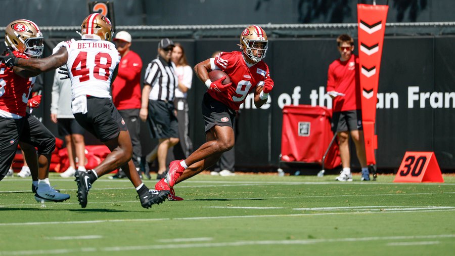 Brayden Willis, in a red 49ers practice jersey with a single-digit 9 and a gold helmet, carrying the ball upfield on a training camp practice field in 2026. Linebacker Tatum Bethune, in a white jersey with 48, pursues from the left. A 30-yard marker stands on the sideline behind them. A practice photograph, not a game.