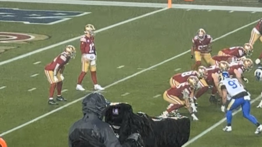 Brock Purdy stands in the shotgun behind his offensive line in the rain at Levi's Stadium, with a Rams defender set across the line of scrimmage and a television camera operator in the foreground. A photograph taken from the stands at a 2024 game, not from this season.