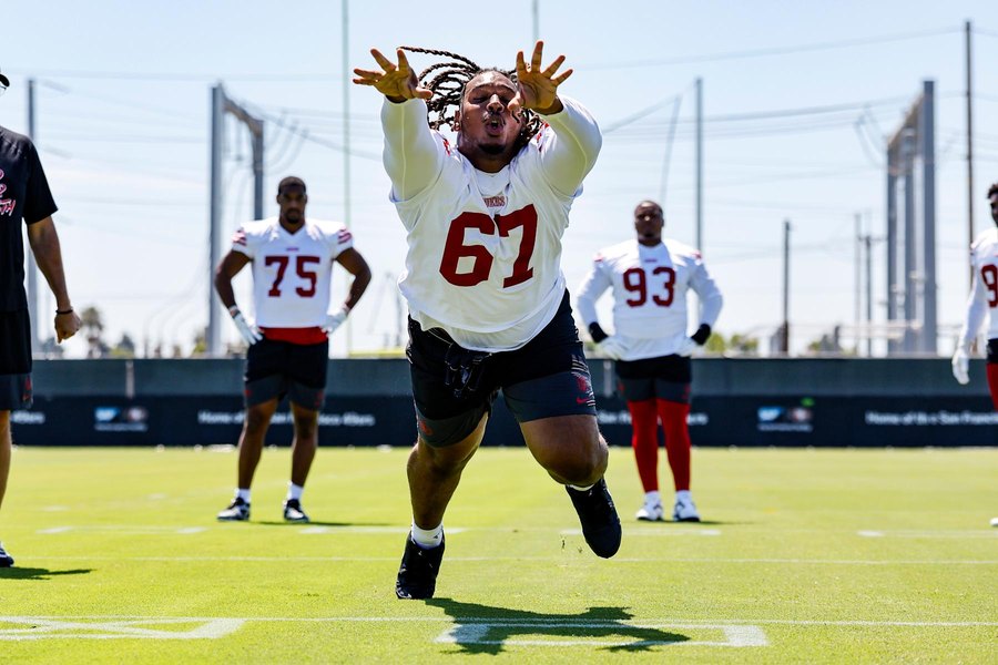Bryson Eason in a white 49ers practice jersey, number 67, airborne with both hands raised to bat down a pass during a drill on the Santa Clara practice fields, a coach at the left edge and two teammates in white practice jerseys standing behind him. This is a camp photograph and not from Thursday's game at the Chargers, in which Eason played 26 defensive snaps.