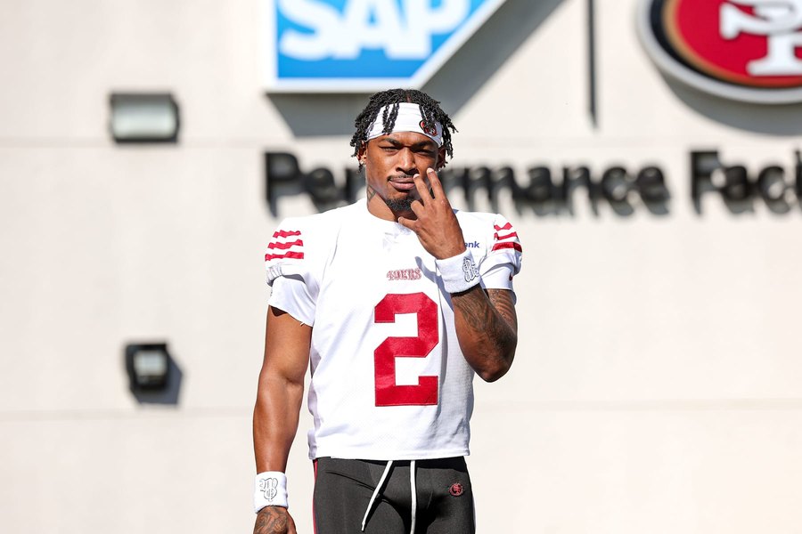 Deommodore Lenoir in the 49ers' white road jersey with the number 2 on his chest, standing outside the team's Santa Clara facility beneath the Performance Facility signage, one hand raised to his face. A club gallery photograph, not from Saturday's practice.