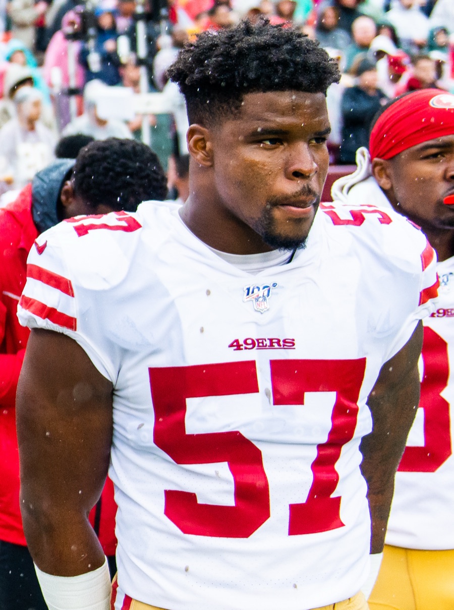 Dre Greenlaw, in the 49ers' white road jersey, stands on the sideline in light rain during the 2019 season, with a crowd in ponchos behind him.