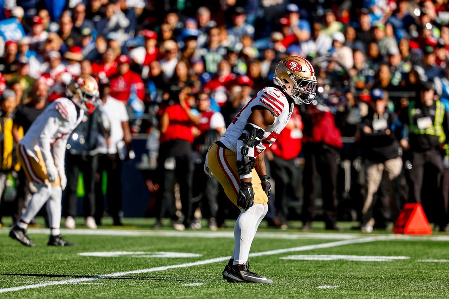 Dre Greenlaw, in the 49ers' white road jersey, gold trousers and gold helmet, stands side-on in a two-point stance at the line at Lumen Field on Oct. 11, 2026, with a teammate out of focus at left and a full crowd behind him.