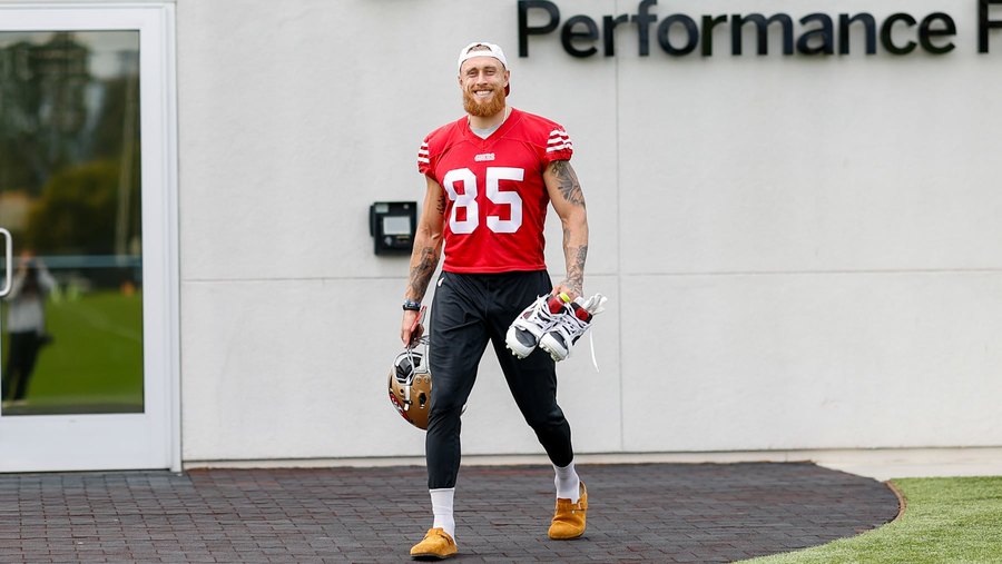 George Kittle walking out of the 49ers' Santa Clara Performance Facility in a red practice jersey numbered 85 and black tights, smiling, carrying his helmet in one hand and his cleats in the other. A club gallery photograph, not from Sunday.