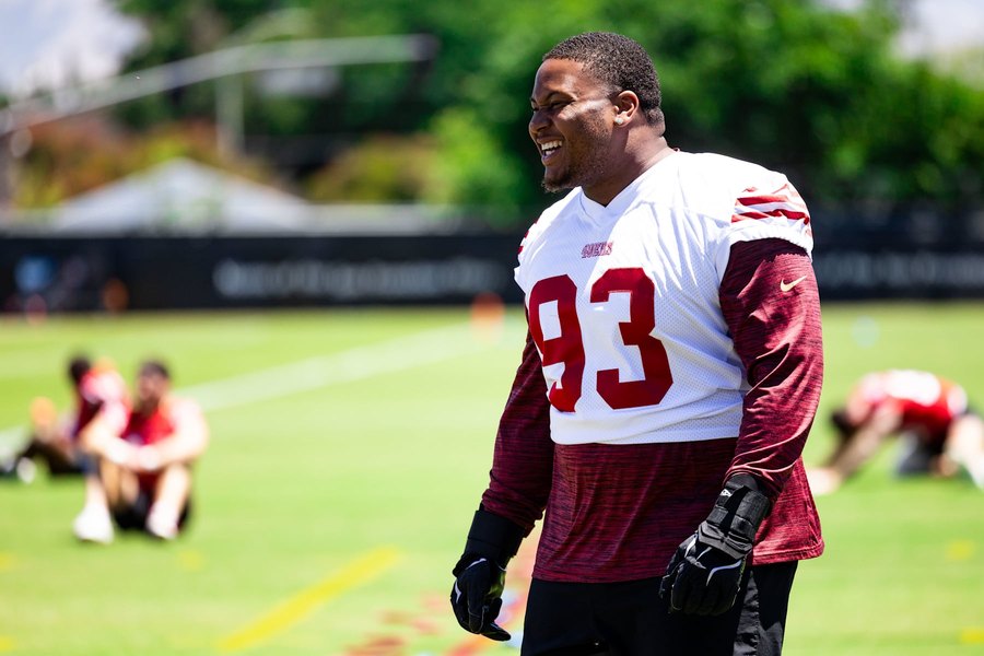 Gracen Halton, number 93, in a 49ers white practice jersey over a red long-sleeve shirt, smiling on the practice field with teammates stretching out of focus behind him. This is a training camp photograph and not from Thursday's preseason game, in which Halton played.