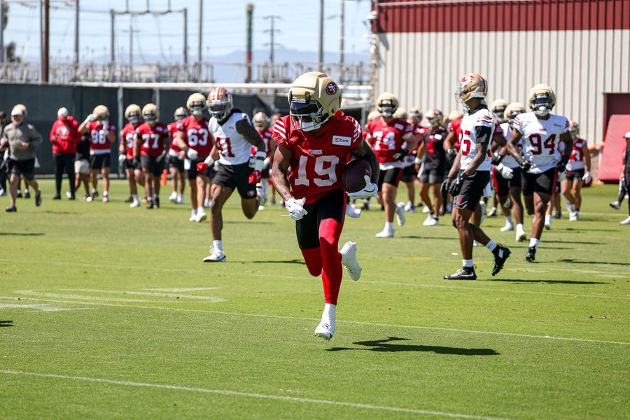 Jacob Cowing running upfield with the ball tucked under his right arm during a 49ers training camp practice in Santa Clara, in a red practice jersey numbered 19, the number he wore before Deebo Samuel signed on August 1 and took it back. A club gallery photograph from July, not from the Chargers game.