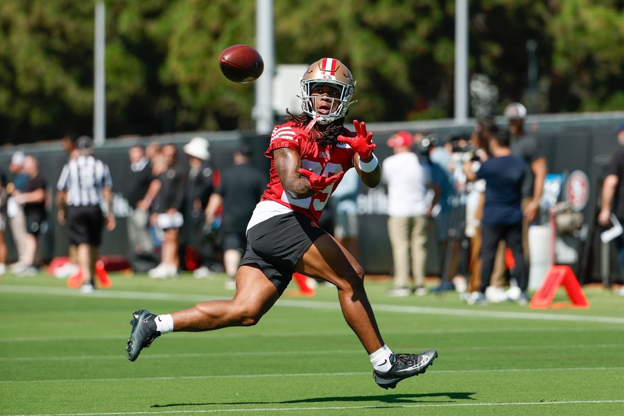 Jordan James, No. 29 on a red 49ers practice jersey and black shorts, running under a pass with both hands raised at a training camp practice in Santa Clara, with media and staff standing behind the sideline barrier. A club gallery photograph, not from Sunday.
