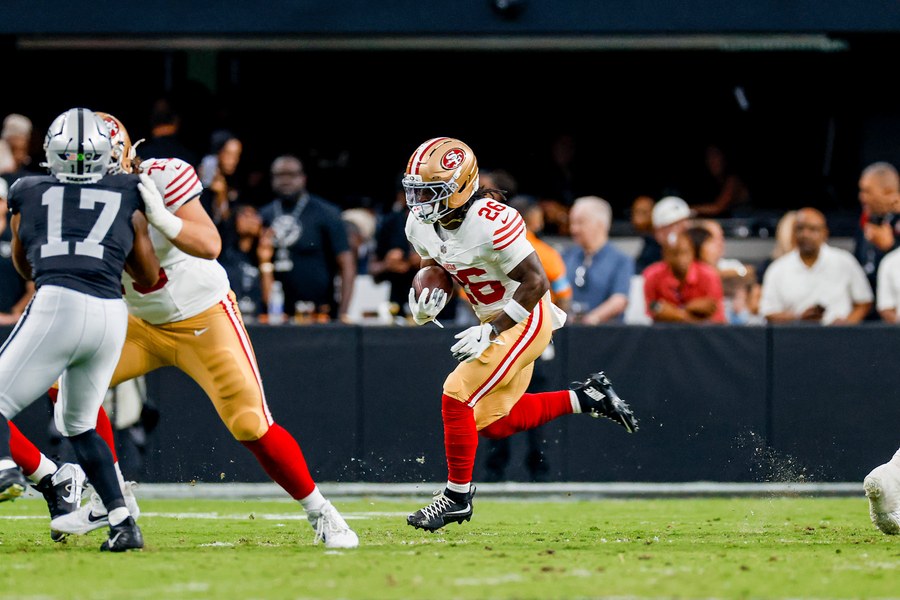 Kaelon Black, in the 49ers' white road jersey with red number 26 and gold trousers, runs upfield with the ball held low in his left hand during the 2026 preseason finale at Las Vegas, a Raiders defender in black and a 49ers lineman engaged at left and fans in the front rows behind him.
