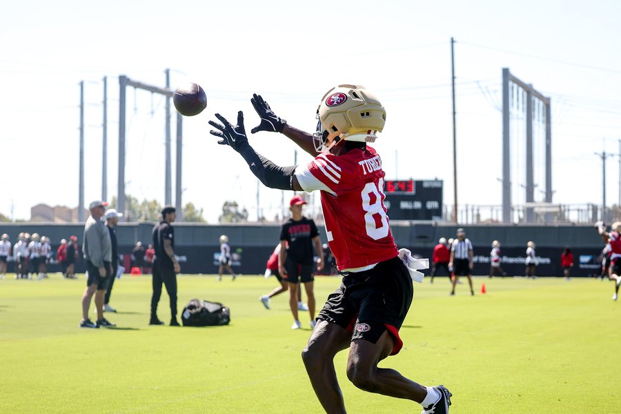 Malik Turner, in the 49ers' red practice jersey with his name on the back and a gold helmet, reaches up with both hands for a ball arriving over his right shoulder on the club's practice field, with staff and players in red and white behind him.