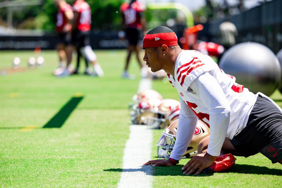 A San Francisco player in the club's white and red practice jersey and a red headband, crouched with one hand on a yard line during a stretching period at the team's Santa Clara facility, gold helmets lined up along the same line beside him and teammates out of focus behind. An offseason practice photograph, not from Thursday's game.