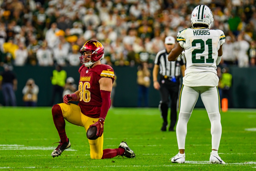 Nate Hobbs in a white Green Bay Packers road uniform during the 2025 season, seen from behind with the nameplate HOBBS and the number 21 legible, a Washington Commanders player in burgundy, number 86, on one knee in front of him and an official in the background. Hobbs wears 22 in San Francisco. This frame is from the last season in which he played a regular-season snap.