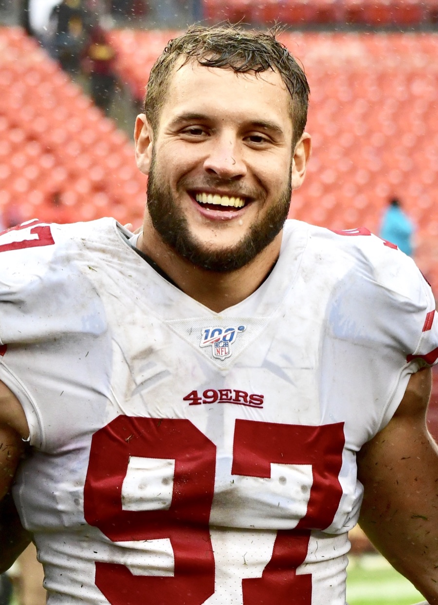 Nick Bosa in the 49ers' white road jersey with red numerals, the 49ERS wordmark across the chest and the number 97 below it, grinning at the camera after a game, the jersey streaked with mud and the stadium seats behind him empty.