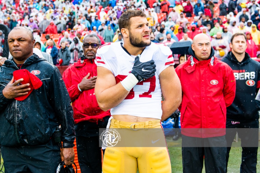 Nick Bosa, bare-headed in a white 49ers road jersey, hand over his heart during the anthem in the rain at Washington in October 2019, with team staff in red jackets beside him. A 2019 photograph, not from this week.