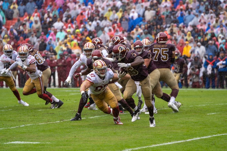 Nick Bosa in the 49ers' white road jersey with red numerals and the number 97 on his chest, gold pants and gold helmet, bent low in the rain as he reaches for a ball carrier in maroon, with Washington's number 75 blocking behind the play.