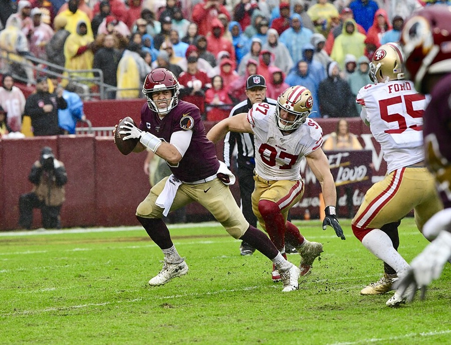 Nick Bosa, in a white 49ers road jersey with 97 in red, gold pants and a gold helmet, closing on a quarterback in a burgundy jersey who is looking downfield with the ball at his ear. Wet grass, rain gear in the stands behind them. A 2019 photograph from a game at Washington, taken seven seasons before the practice week described here.