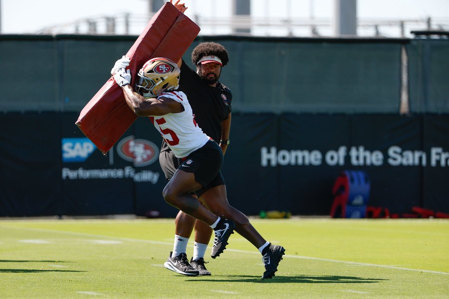 A 49ers player in a white practice jersey and gold helmet drives a red blocking pad off his shoulder on a practice field at the club's Santa Clara facility, a coach in a black shirt and visor a step behind him. The number on his chest is hidden behind the pad and the man is not identified here.