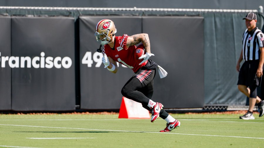 Ricky Pearsall, number 1, in a red 49ers practice jersey and black pants, running upfield with the ball tucked under his right arm during a training camp practice at the SAP Performance Facility, an official jogging behind him and the club's own San Francisco 49ers backdrop along the fence. Pearsall carries the largest injured-reserve charge on either money book at $3,419,563.