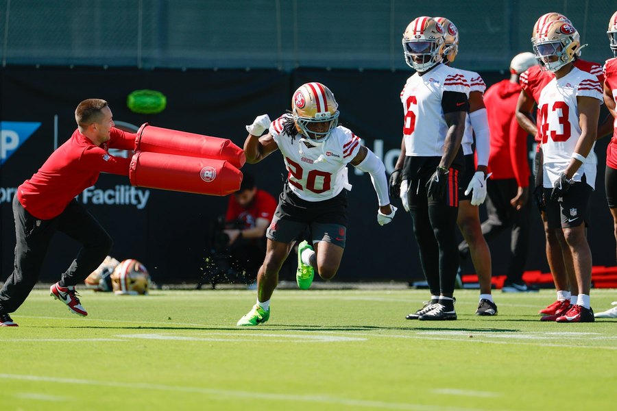 Upton Stout bursting past a coach holding a padded shield during a 49ers training camp drill in Santa Clara, in a white practice jersey numbered 20, with teammates waiting their turn behind him. A club gallery photograph, not from Sunday's practice.
