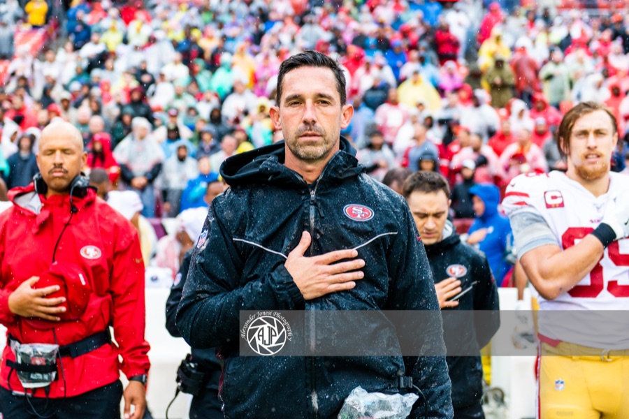 Kyle Shanahan in a black 49ers hooded jacket, hand over his heart during the anthem in heavy rain, with George Kittle in the white 49ers road jersey behind him. This is a 2019 road-game photograph and not from Tuesday's joint practice in El Segundo.
