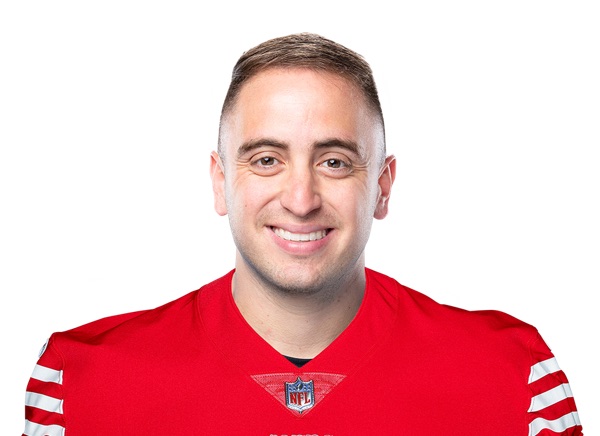 Eddy Piñeiro photographed from the chest up against a plain white background, smiling into the camera, short dark hair, wearing a red 49ers jersey with white and red striped sleeve trim and the NFL shield at the collar. A posed league headshot, not football action.