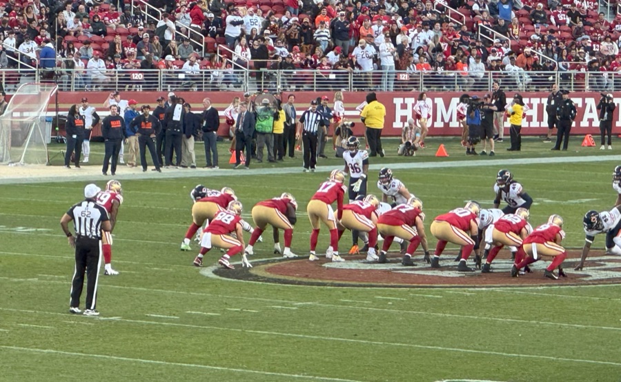 The 49ers offense set at the line of scrimmage in home red at Levi's Stadium, the offensive line in a three-point stance across the middle of the frame, a Chicago defense across from them and an official standing at the left sideline.