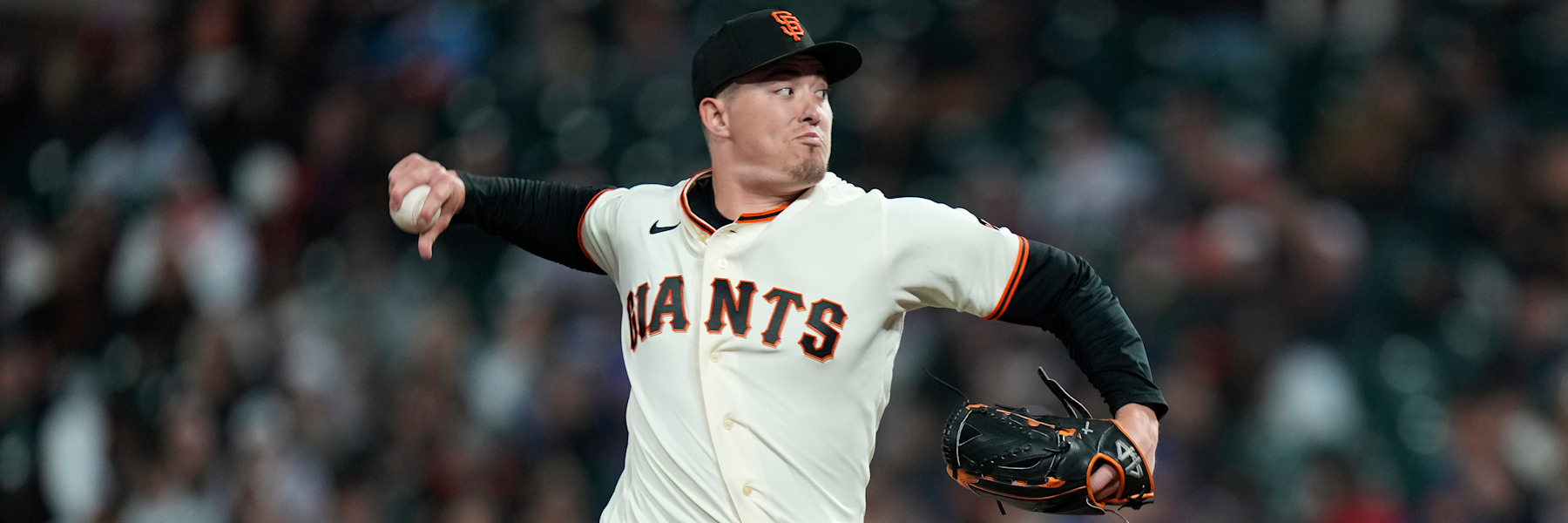 Blade Tidwell in the Giants' cream home uniform, mid-delivery with the ball still in his right hand and his glove trailing, a blurred ballpark crowd behind him.