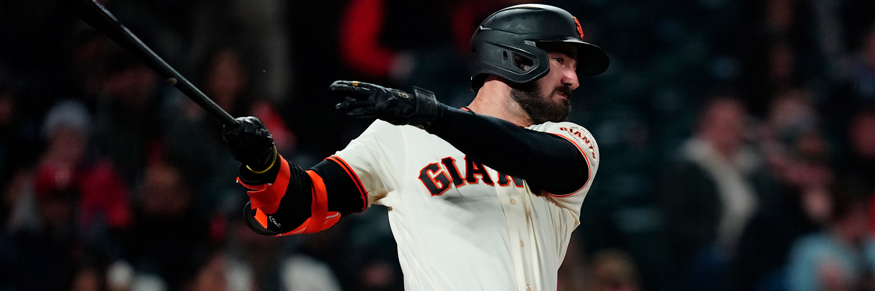 Bryce Eldridge at the end of a swing in San Francisco's cream home uniform, the black and orange GIANTS script across his chest, a black helmet with the orange interlocking SF, his top hand off the bat and his right arm extended, his head turned to follow the ball. A blurred crowd behind him. A league media photograph, not from Tuesday's game.