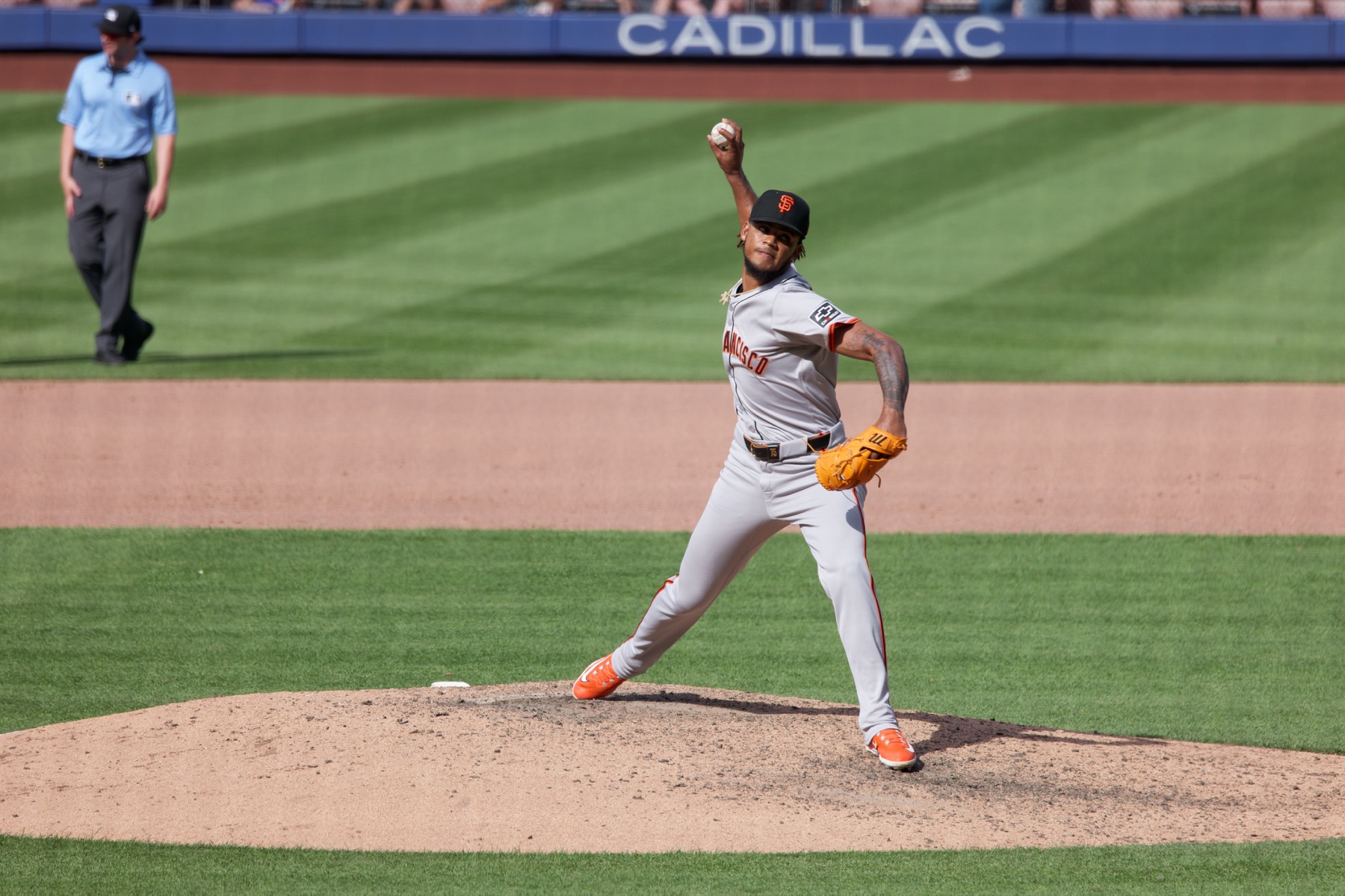 Camilo Doval in the Giants' gray road uniform, black SF cap and orange cleats, striding toward the plate from the mound during a day game in May 2024, an umpire behind him in the outfield grass.