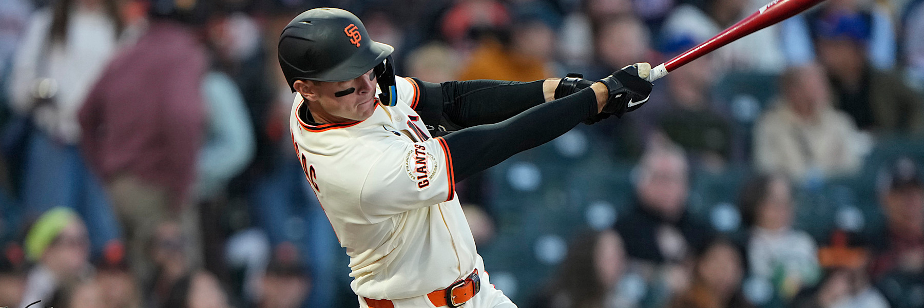Daniel Susac finishing a swing in the Giants' cream home uniform, GIANTS scripted across the chest in black and orange, black batting helmet with the orange SF, a blurred Oracle Park crowd behind him.