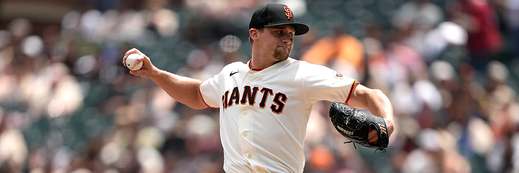 Keaton Winn in mid-delivery in San Francisco's cream home uniform, the GIANTS wordmark in black and orange across his chest, a black cap with the orange interlocking SF, the ball in his raised right hand and his glove extended out to the side. A blurred crowd behind him. A league media photograph, not from Thursday.