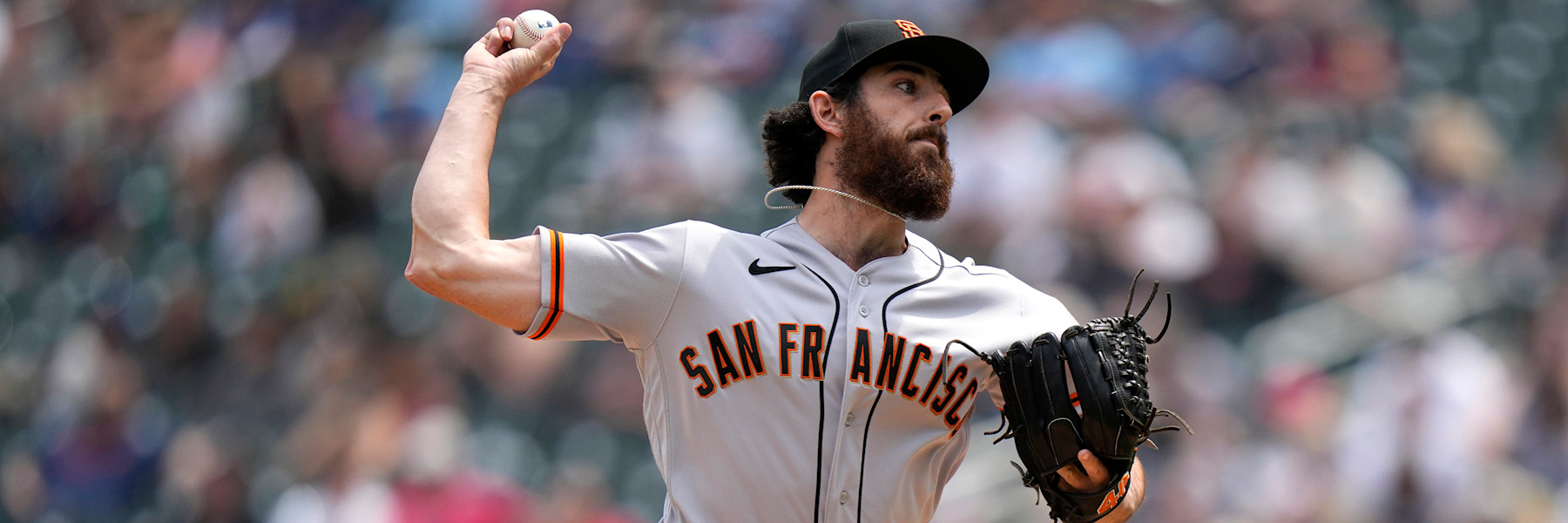 Ryan Walker mid-delivery in San Francisco's grey road uniform, the SAN FRANCISCO wordmark in black and orange across his chest, a black cap with the orange interlocking SF, the ball held up behind his head in his right hand and his glove out in front of him. A blurred crowd behind. A league media photograph, not from Saturday.