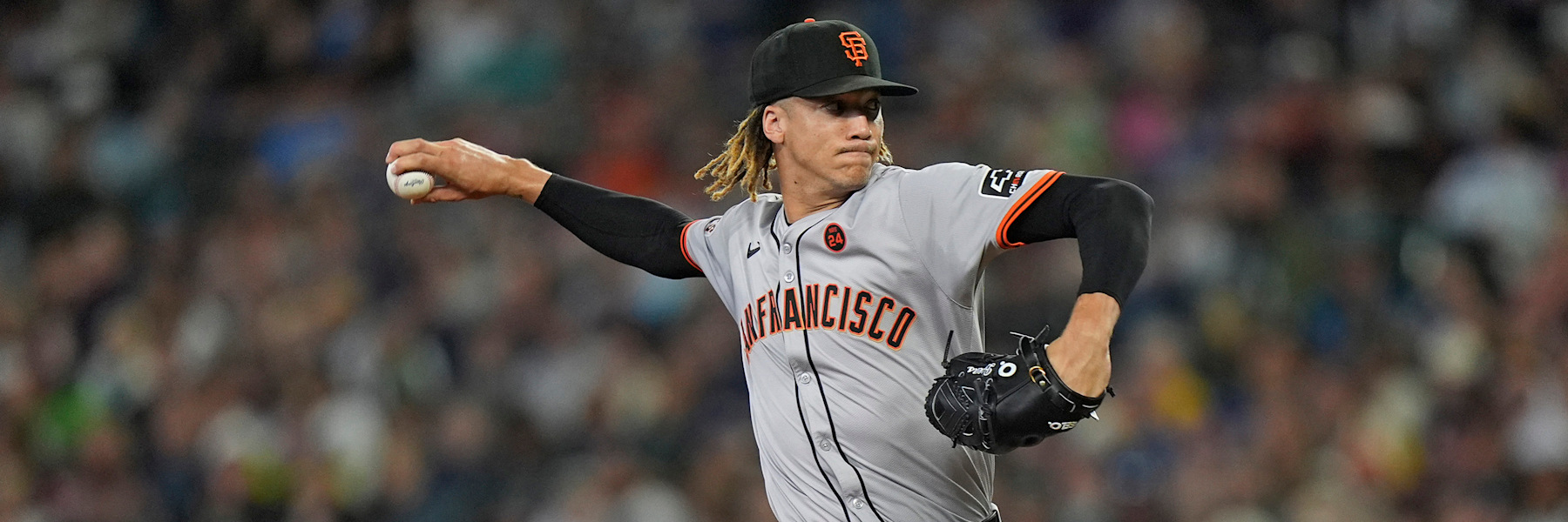 Spencer Bivens at the release point for San Francisco, right arm cocked back with the ball visible in his hand and his black glove tucked in front of his chest, wearing the Giants grey road uniform with SAN FRANCISCO in orange script, a black cap with the orange interlocking SF, and the circular 24 memorial patch on the right chest of the jersey. Dreadlocks under the cap, a blurred crowd behind him. The date of the photograph is not established.