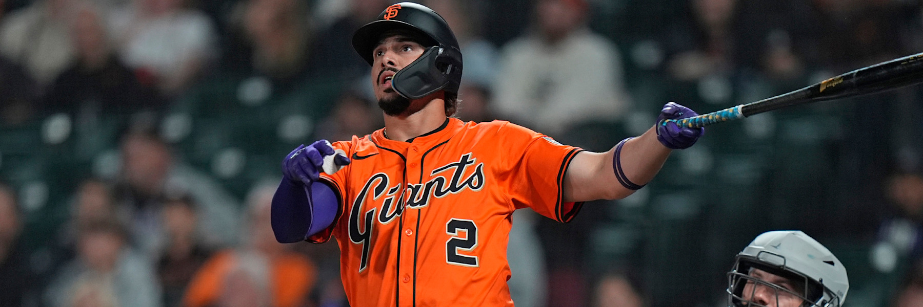 Willy Adames of the San Francisco Giants at the plate, mid-follow-through, watching the ball after a swing. He wears the Giants orange alternate jersey with the black-outlined script and the number 2, and a black helmet with the orange SF. A catcher's mask is visible at the right edge and the crowd behind him is out of focus.