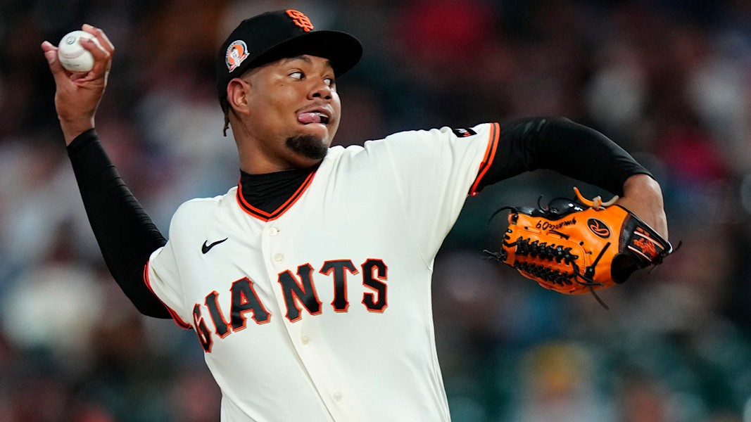 Yunior Marte at the top of his delivery for San Francisco, the ball in his raised right hand and his orange glove out in front of him, in the Giants home cream jersey with the black and orange GIANTS script and a black cap with the orange interlocking SF, the stands at Oracle Park out of focus behind him. The date of the photograph is not established, so it is not from Friday night.
