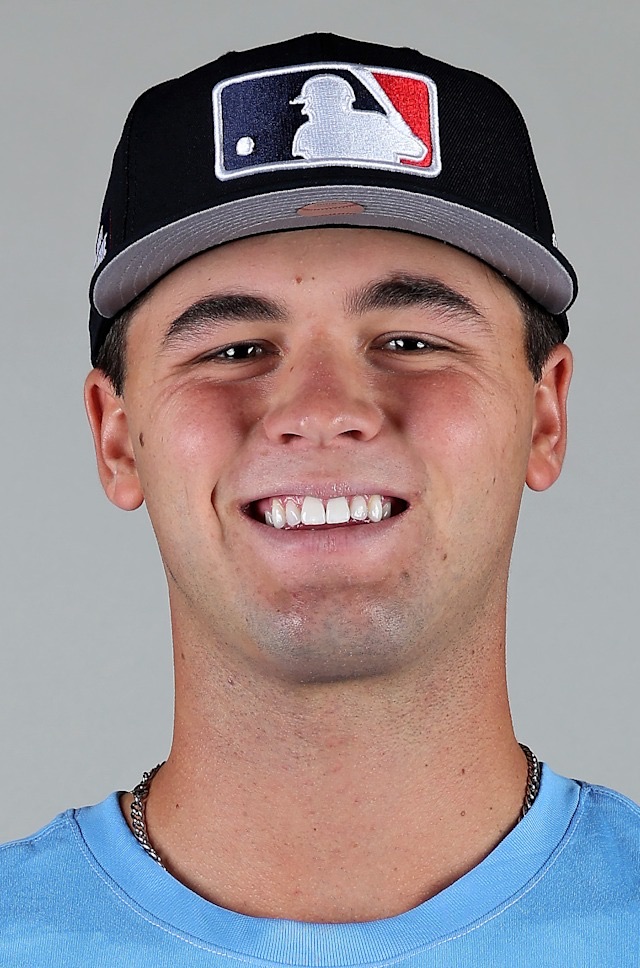 Jackson Flora smiling in a posed head-and-shoulders portrait against a plain grey background, wearing a black cap with a grey brim carrying the Major League Baseball silhouette logo and a light blue shirt. Not a Giants uniform. A league media photograph, not from Friday night.