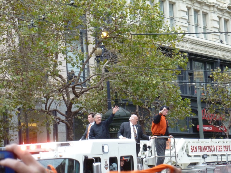 Four Giants broadcasters ride atop a San Francisco Fire Department ladder truck on Market Street during the 2012 World Series victory parade, two of them waving to a crowd out of frame.