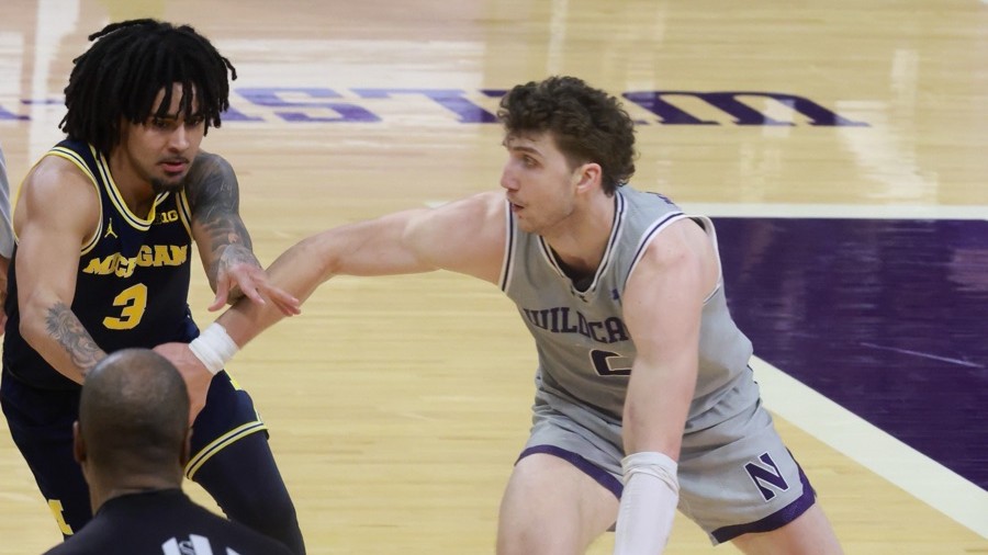Elliot Cadeau, in Michigan's navy road jersey, guards a Northwestern player in gray who has an arm stretched toward him, on the hardwood and purple floor at Northwestern on February 11, 2026.