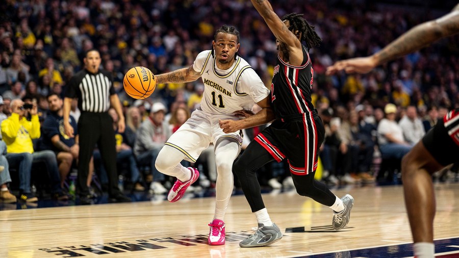 Roddy Gayle Jr. in Michigan's white uniform drives left-handed past a Houston defender in black and red during the exhibition in Grand Rapids in October 2026, with a referee and the crowd behind them.