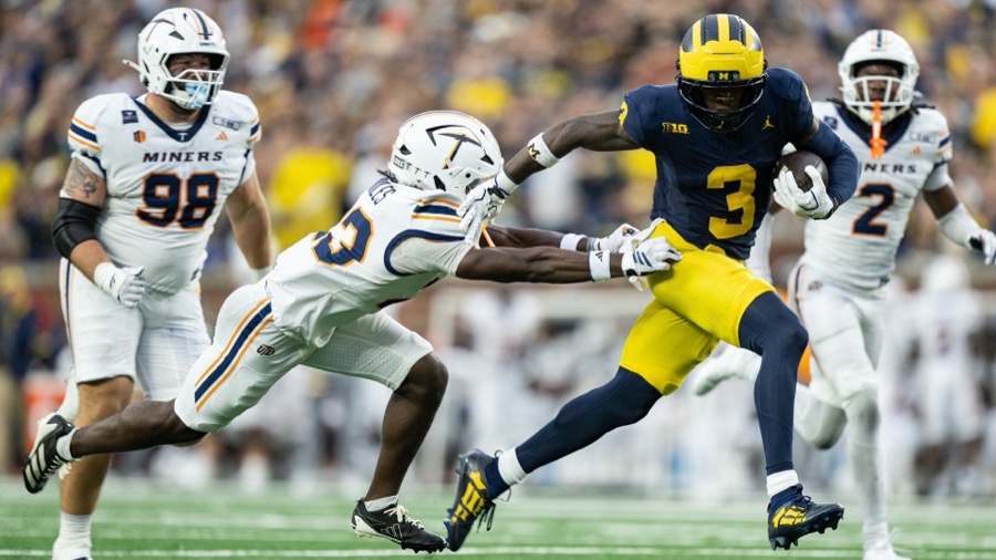 Andrew Marsh, number 3 in Michigan's navy home jersey and maize trousers, carries the ball in his left arm as a UTEP defender dives at him at Michigan Stadium on September 19, 2026, a week before the Iowa game.