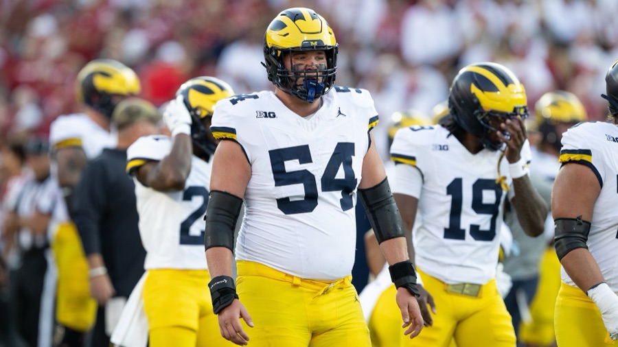 Andrew Sprague on the field at Oklahoma in September 2025, standing between snaps with his arms down and teammates around him, in the Michigan road white with maize trousers and the winged helmet, number 54. A crimson Oklahoma crowd fills the stands behind him. The photograph is from the 2025 season and is not from this year.
