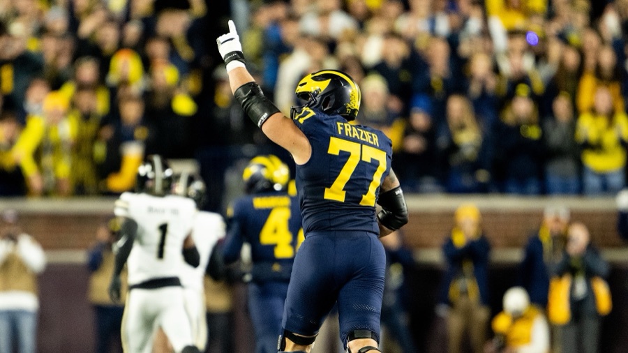 Blake Frazier, number 77 in Michigan's navy home jersey, seen from behind with his left arm raised and one finger pointing up during the Purdue game at Michigan Stadium in 2025, the crowd behind him in maize.