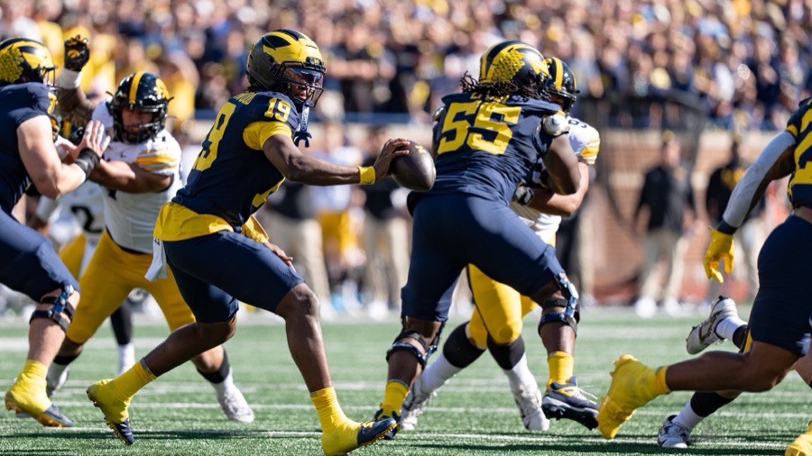 Bryce Underwood, number 19 in Michigan's navy home jersey and maize trousers, holds the ball out at his side behind a block against Iowa at Michigan Stadium on September 26, 2026, Iowa's defenders in white.