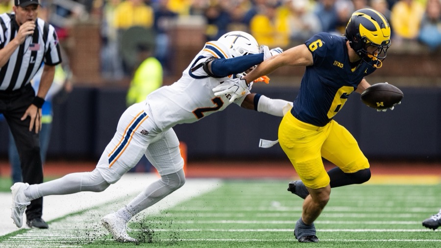 JJ Buchanan, number 6 in Michigan's navy home jersey and maize trousers, carries the ball in his right hand and stiff-arms a UTEP defender along the sideline at Michigan Stadium on September 19, 2026, with an official watching from the left.