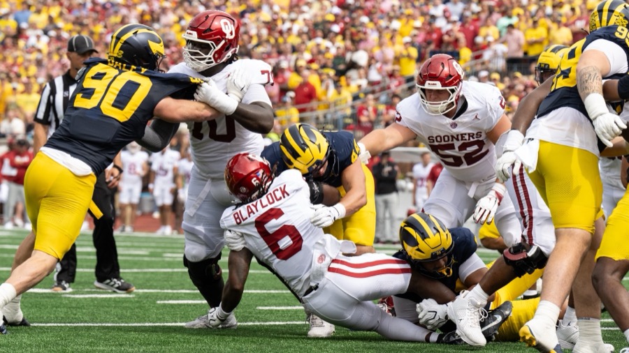 John Henry Daley, number 90 in Michigan's navy home jersey, engaged with an Oklahoma lineman while teammates bring down Oklahoma's number 6 at the line during the September 12, 2026 game at Michigan Stadium.