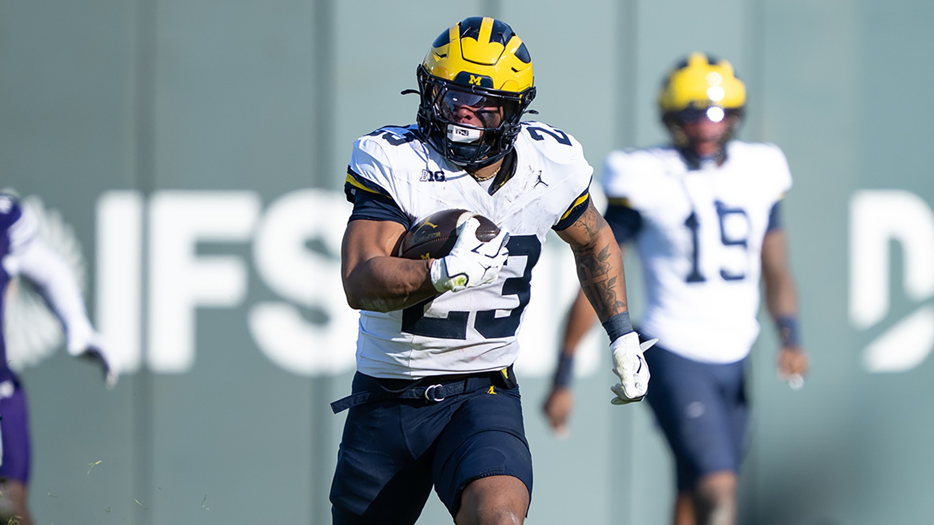 Jordan Marshall, number 23 in Michigan's white road jersey and navy pants, tucking the ball under his right arm and running to his left at Northwestern in 2025. A teammate wearing 19 jogs in the background and a Northwestern player in purple is out of frame to the left.