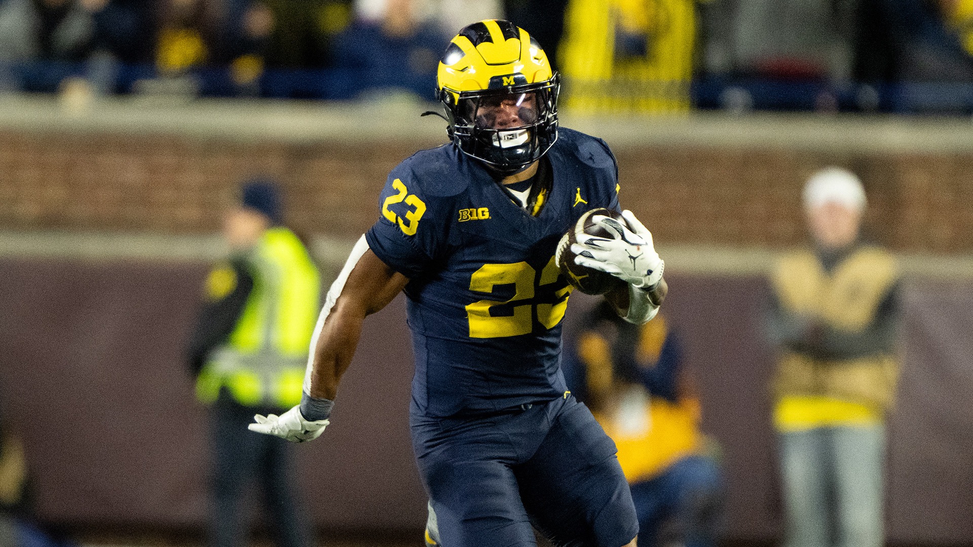 Jordan Marshall, number 23 in Michigan's navy home jersey with maize numerals and a maize-and-navy winged helmet, carrying the ball in his right hand and running upfield under the lights at Michigan Stadium in 2025. Two staff members in high-visibility vests are blurred behind him on the sideline.