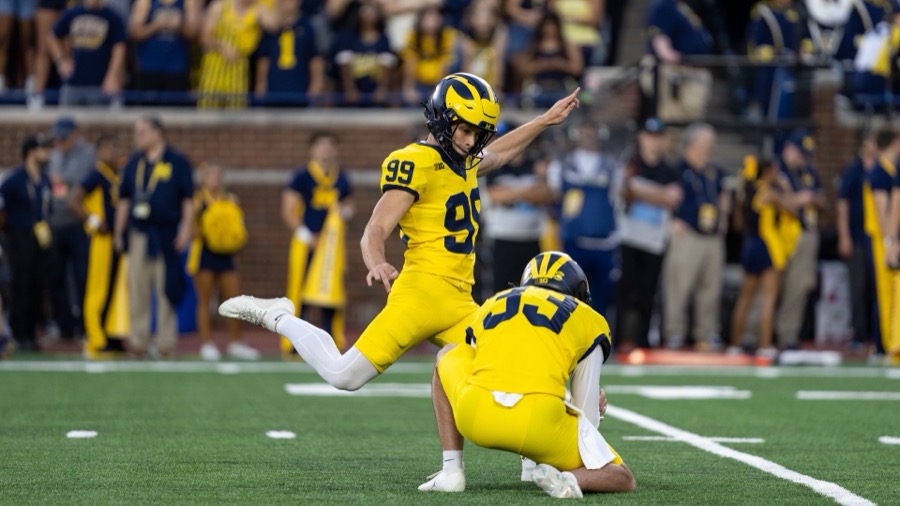 Trey Butkowski, number 99 in Michigan's all-maize alternate uniform and winged helmet, points his left arm forward as he follows through on a kick held by Cam Brown, number 33, kneeling at right, at Michigan Stadium during the September 5, 2026 opener against Western Michigan.