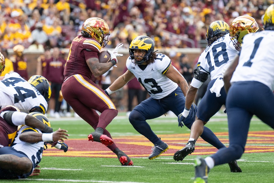 Michigan defenders in white road jerseys, navy trousers and winged helmets close on a Minnesota ball carrier in maroon at Huntington Bank Stadium in Minneapolis on October 3, 2026: linebacker Aisea Moa, number 13, squared up in front of him, Troy Bowles, number 18, seen from behind at right, and number 44 at left.