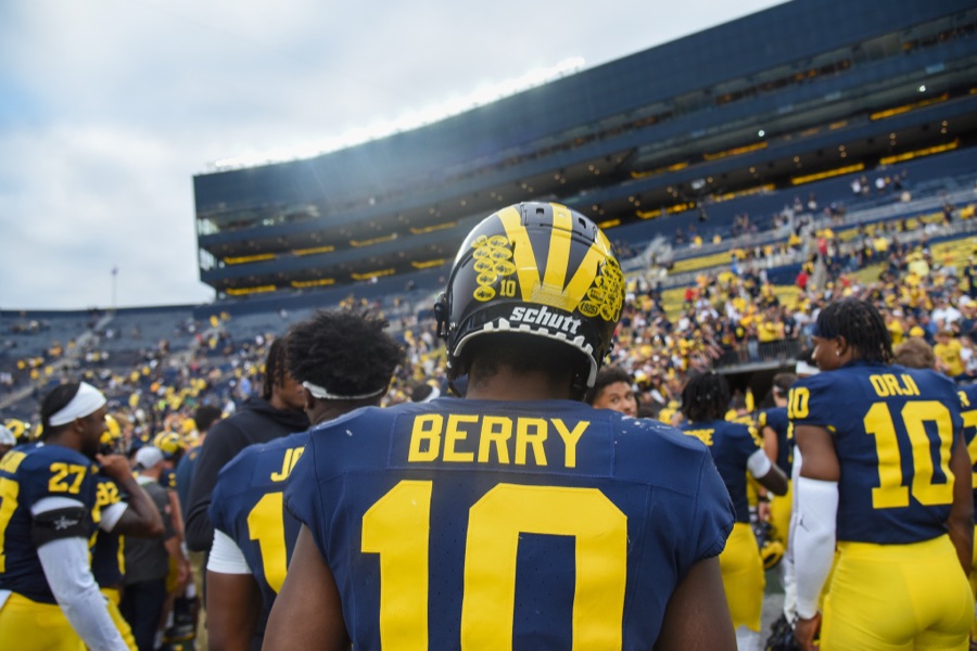 Zeke Berry, shot from behind in Michigan's navy home jersey with BERRY and a maize 10 across the shoulders, stands among teammates and staff on the Michigan Stadium field at Michigan's home game against UNLV in September 2023, the stands behind him only partly filled.