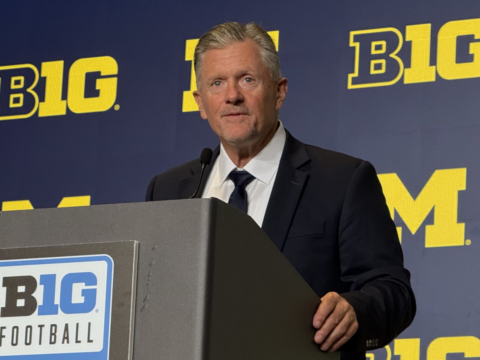 Kyle Whittingham, white-haired and in a navy suit and navy tie, speaking at a lectern carrying the Big Ten Football mark, against a navy backdrop repeating the Big Ten wordmark and a maize block M.